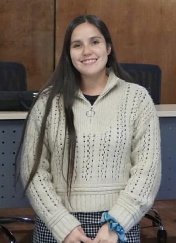 Young woman with long dark hair in a cream knitted sweater, smiling, seated with hands folded in her lap.