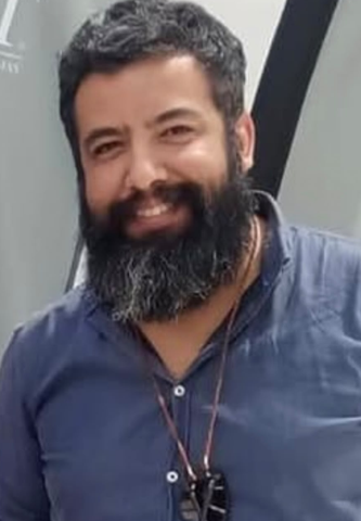Smiling man with a thick black beard wearing a blue button-up shirt to the chest level.