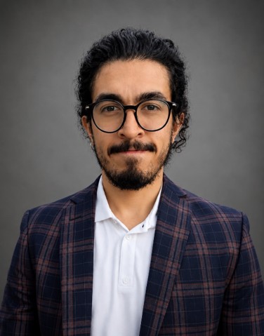 Professional headshot of a man with dark hair and a beard, wearing round glasses, a plaid blazer, and a white shirt against a gray backdrop.