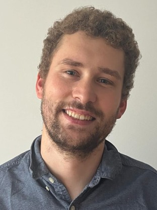 Portrait of a smiling man with curly hair and a beard wearing a blue button-up shirt.
